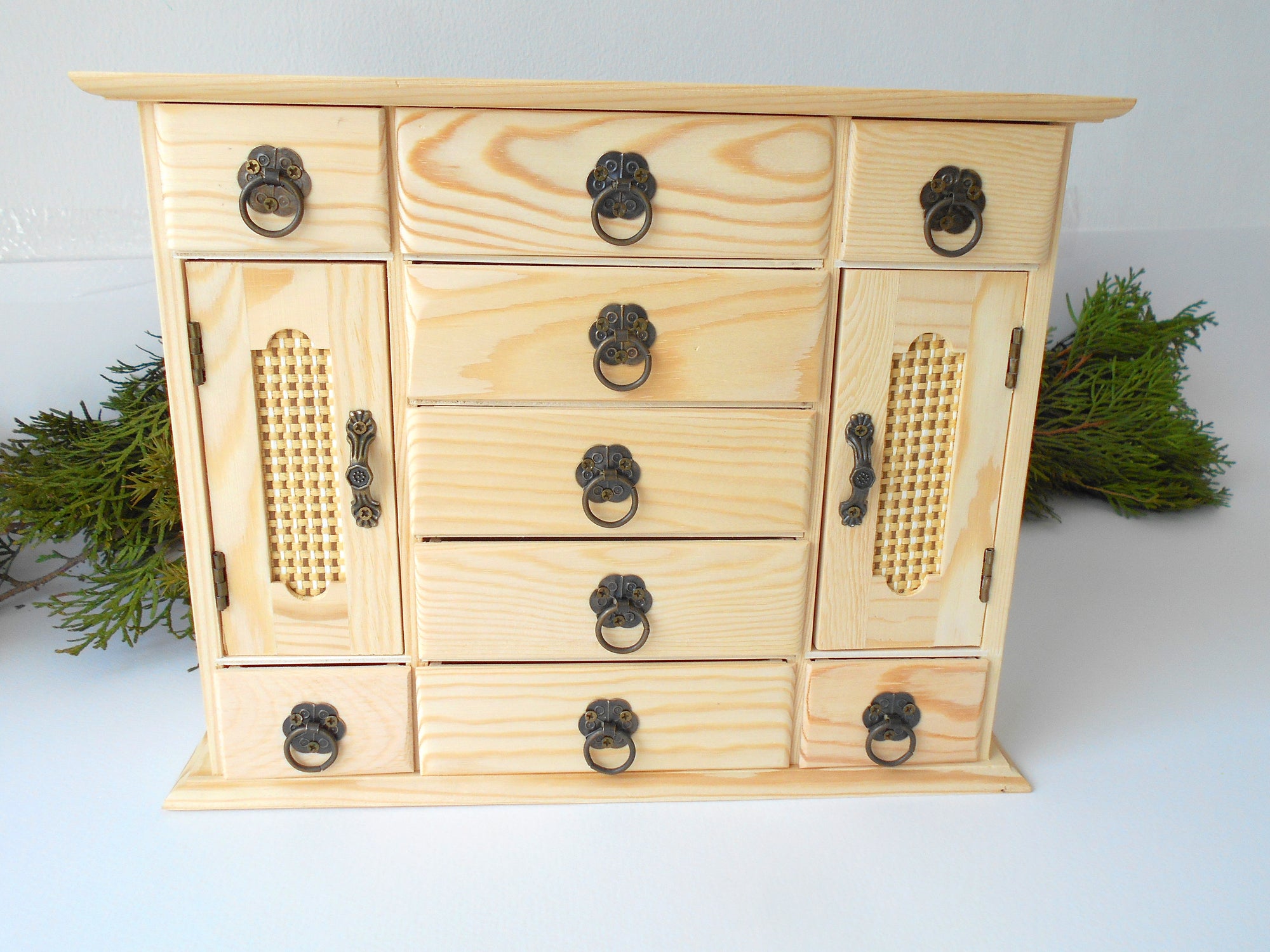 This wooden box with 9 drawers and 2 wardrobe doors is made of pinewood on the outside and bamboo wood for the inside of the drawers. It has metal pulls with a vintage bronze color. The surface of the box is smooth and refined with sanding paper. The color of the box is plain pinewood color.&nbsp;