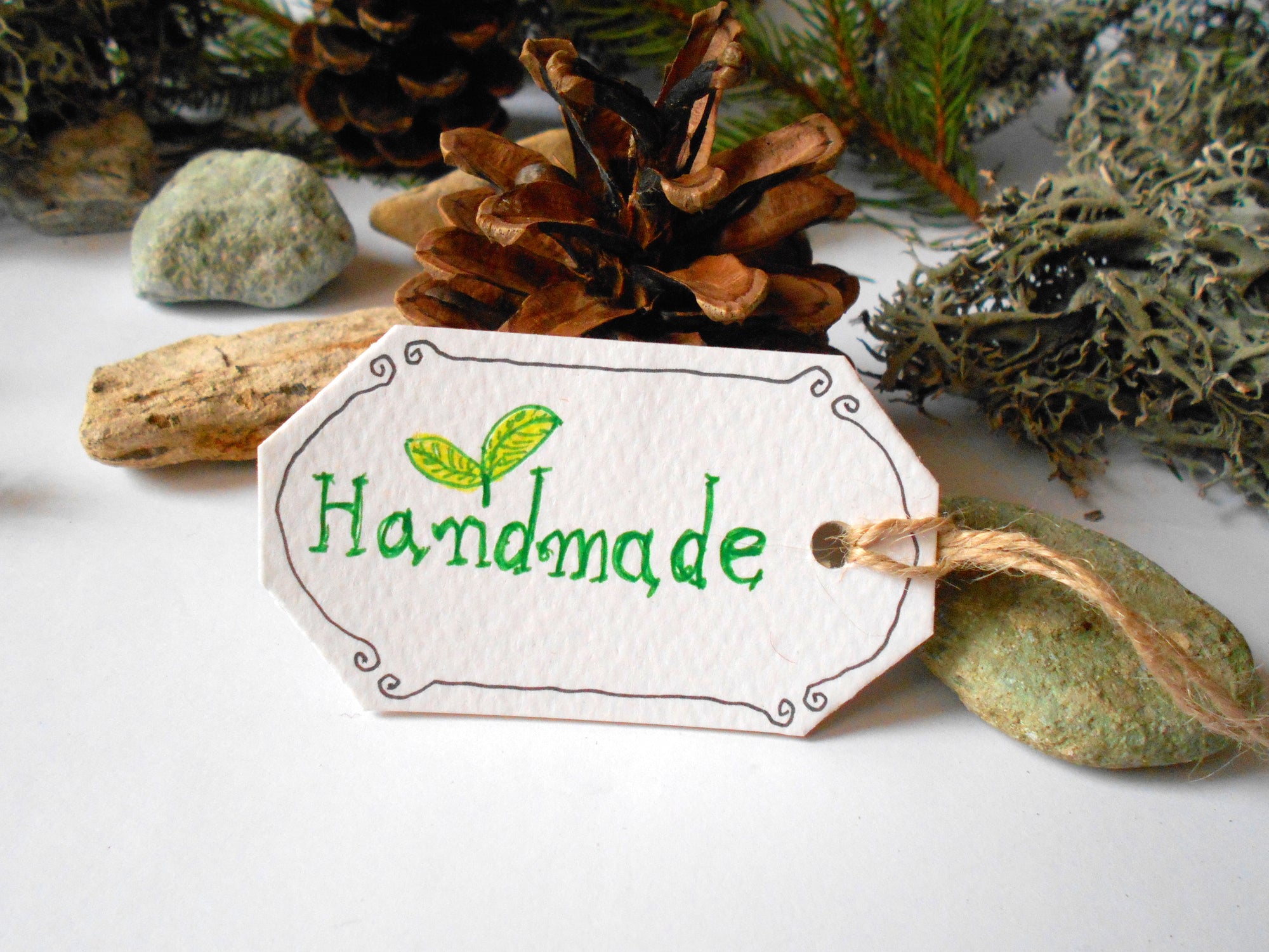 Handmade hang tag with hand-written handmade word on it and surrounded by a pine branch, pine cone, stones and lichen 