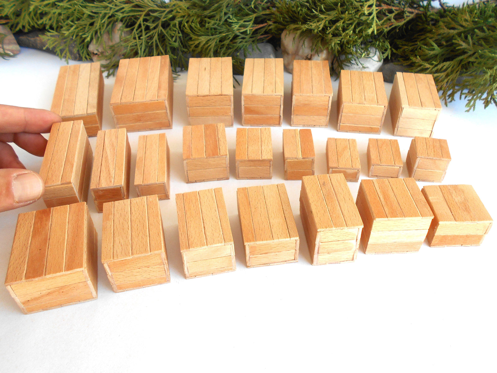 
          
            A set of small natural wood miniature boxes or tiles arranged on a white surface, with a hand holding one block on the left and evergreen branches in the background.
          
        