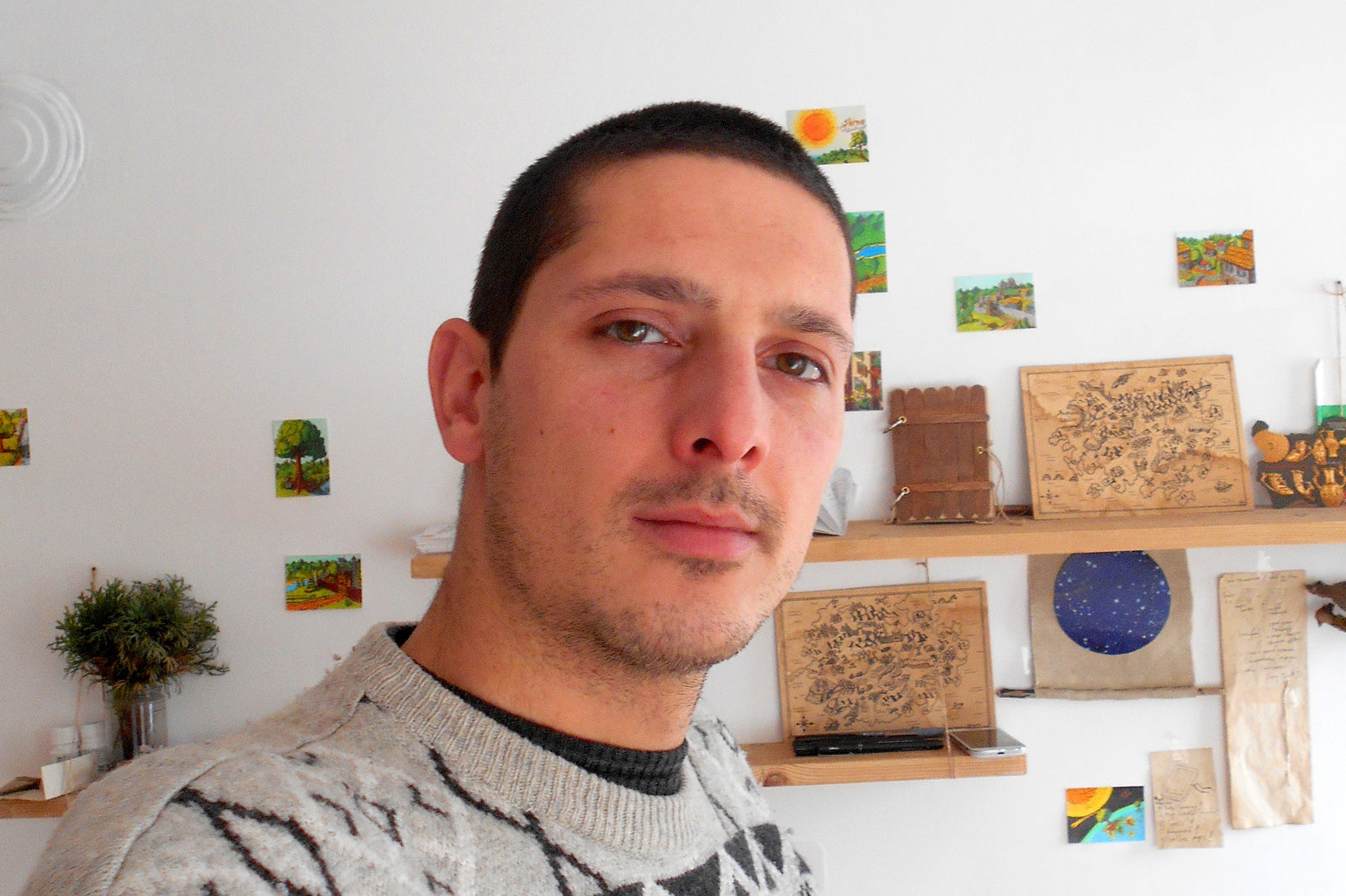 Artist Hristo Hvoynev watching towards the camera and behind him are his shelves with crafts, maps, art prints 