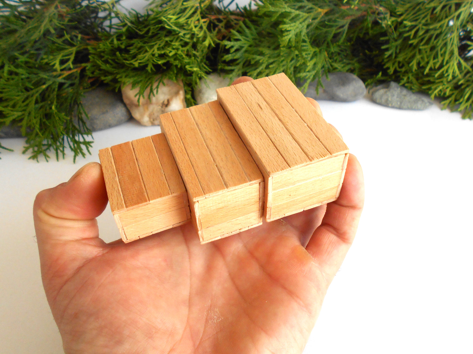 Three miniature wooden blocks with a plank pattern, held in a hand to show scale, with green pine branches and small stones in the background.