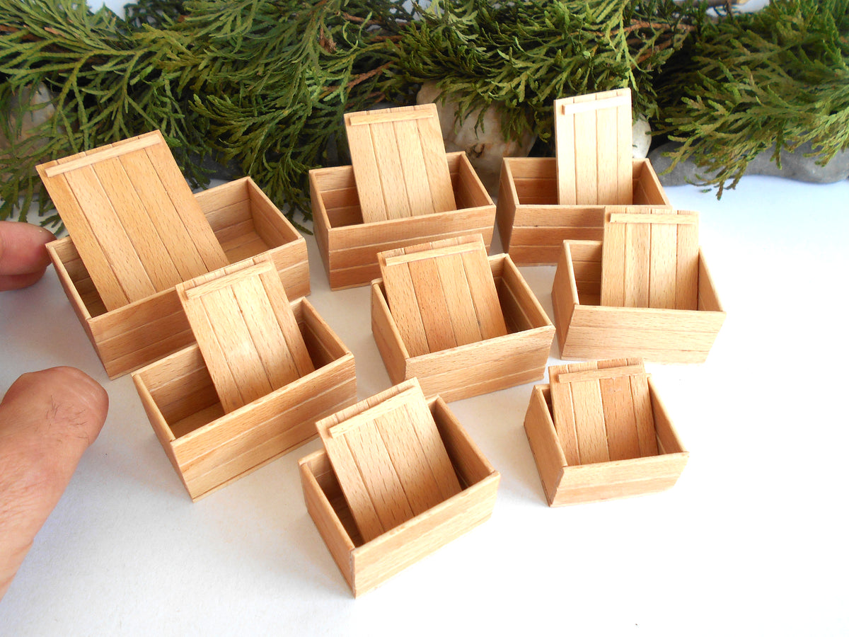 A set of miniature light wood boxes with slatted sides, shown on a white surface with green foliage in the background. A person's fingers are visible on the left edge holding one of the boxes.