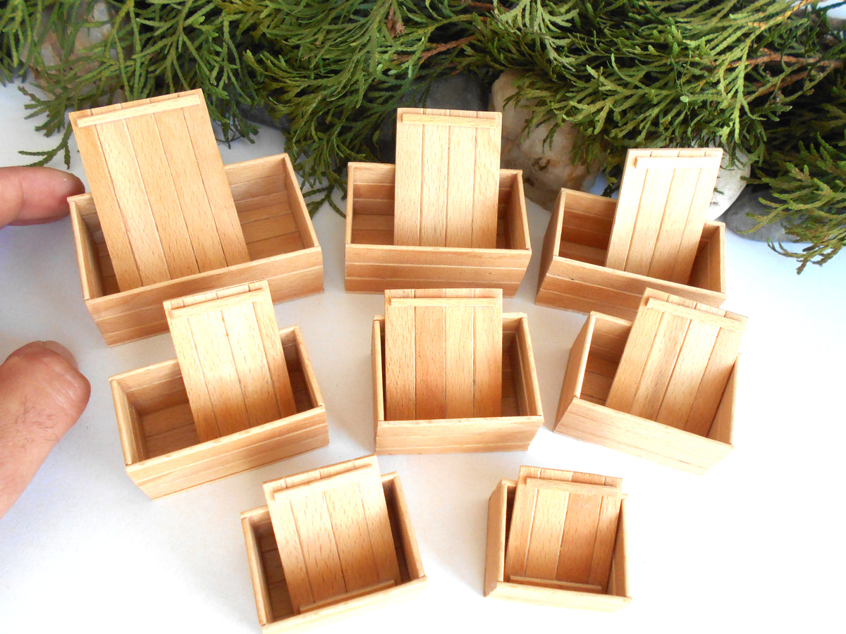A set of miniature light wood boxes with slatted sides, arranged on a white surface with green foliage in the background. Two fingers are visible at the left edge holding one of the boxes.