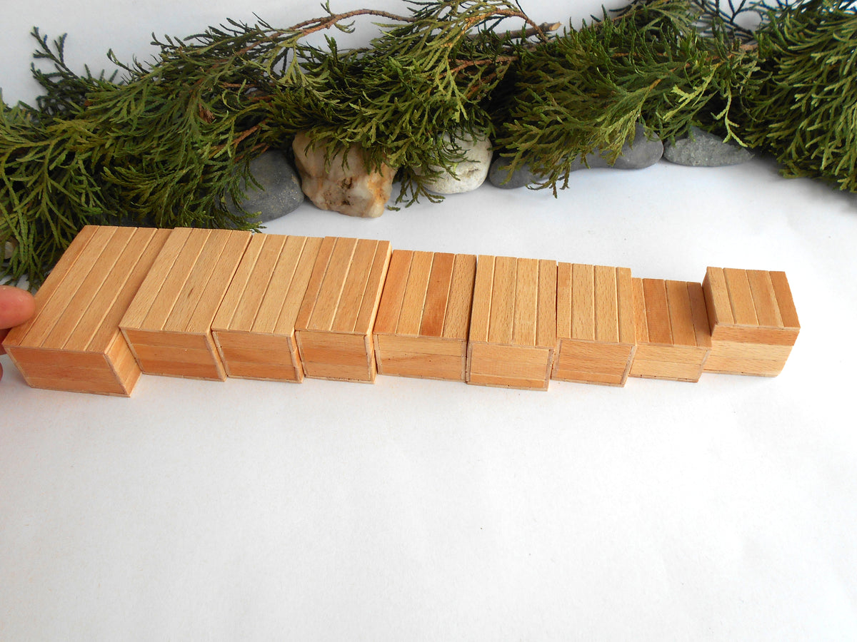 A set of natural light wood mini boxes arranged in a stepped row on a white surface, with green foliage and small stones in the background.