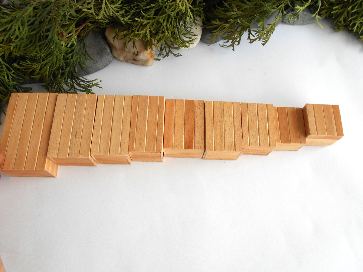A set of wooden Montessori-like mini boxes arranged in a descending size sequence on a white surface, with green foliage and stones partially visible at the top edge.