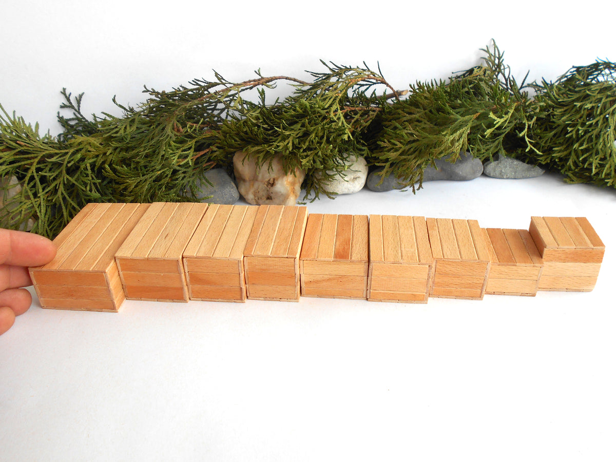A hand holds a row of natural light wood rectangular mini boxes or tiles on a white surface, with green garland and small stones in the background.