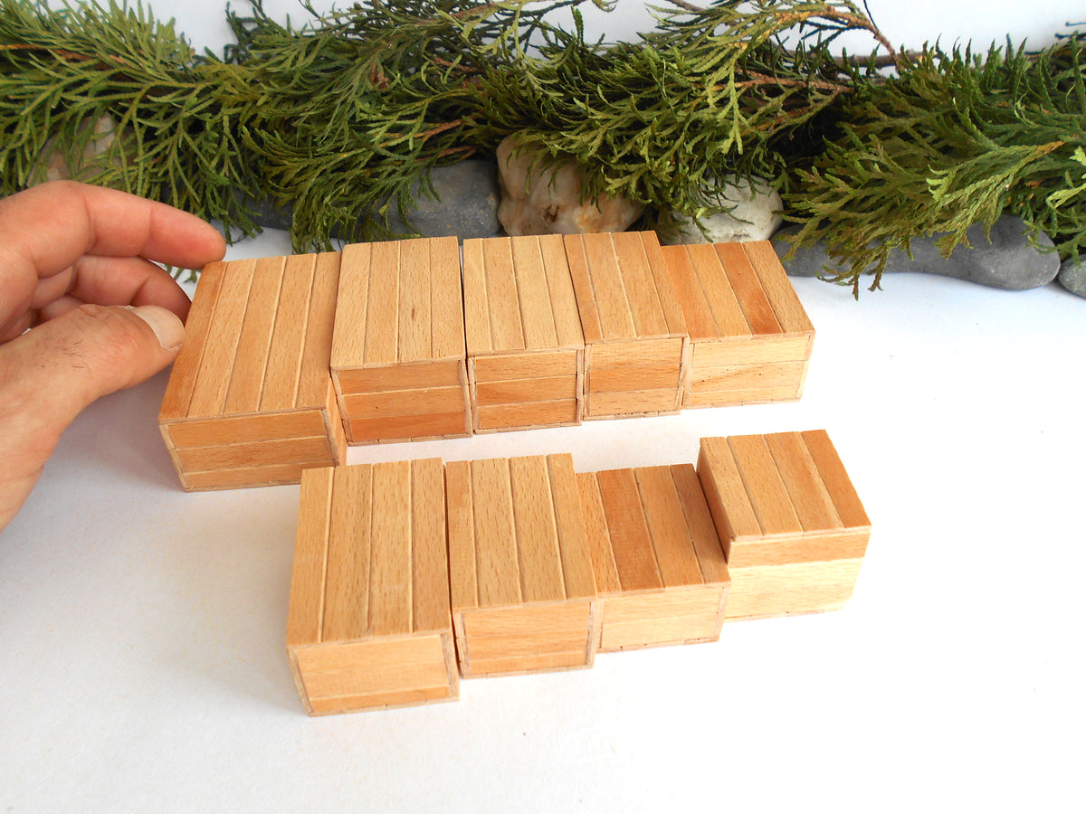 A hand holding two small wooden mini boxes sets on a white surface, with green foliage and gray stones in the background.