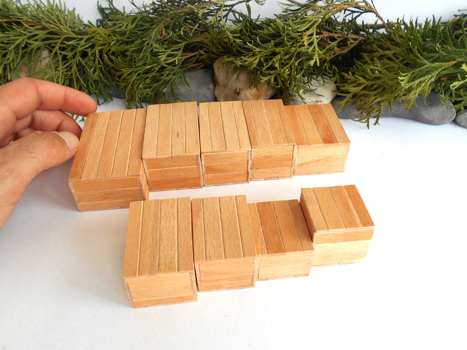 A hand holding two small wooden mini boxes sets on a white surface, with green foliage and gray stones in the background.
