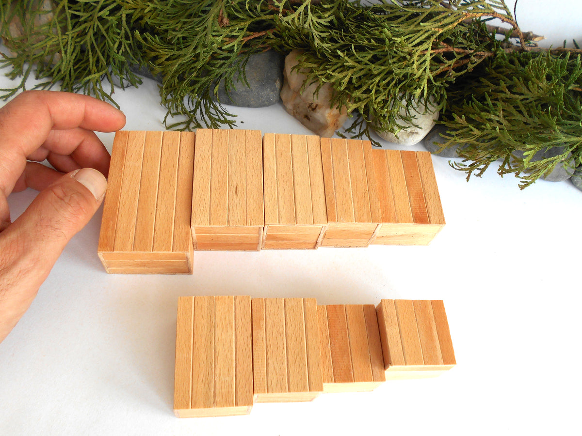 A hand holding a set of light natural wood rectangular miniature boxes , with a second matching set below on a white surface, styled with green foliage and stones in the background.
