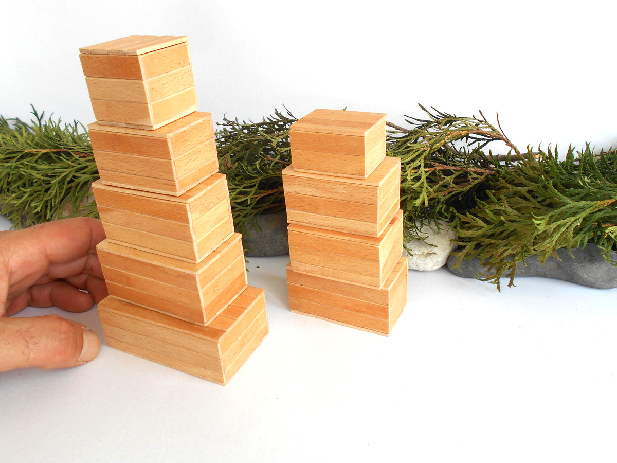 Two stacks of small natural wood miniature boxes on a white surface, with a hand holding the left stack and greenery in the background.