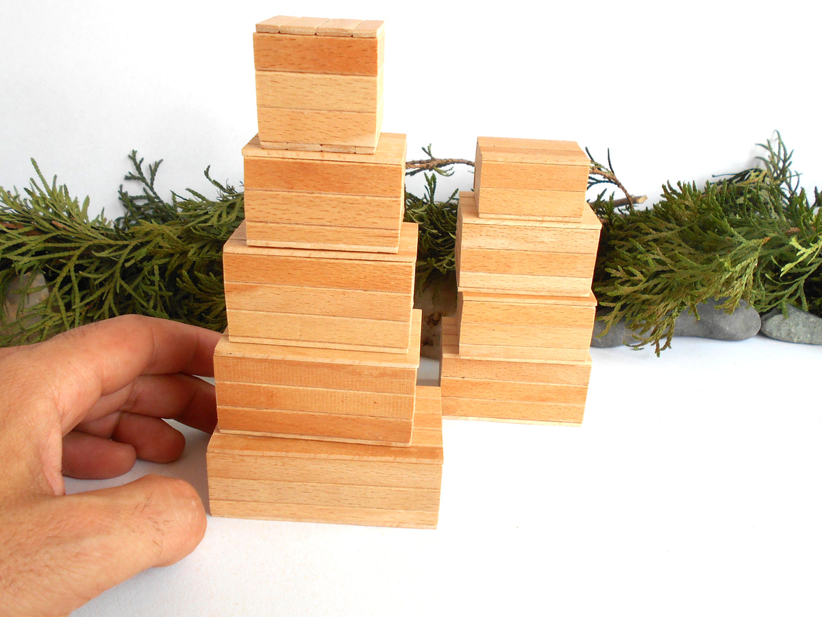 A hand is holding a stack of light natural wood miniature boxes next to two pre-assembled wooden boxes towers, with green foliage and small stones in the background against a white surface.