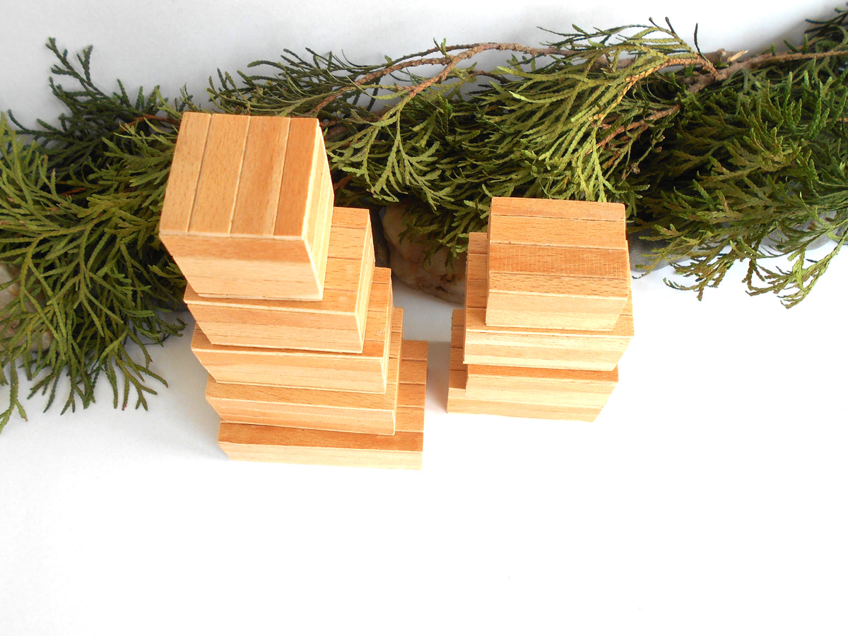 Two stacks of small natural wood miniature boxes , one taller on the left and one shorter on the right, arranged on a white surface with green pine branches behind them.