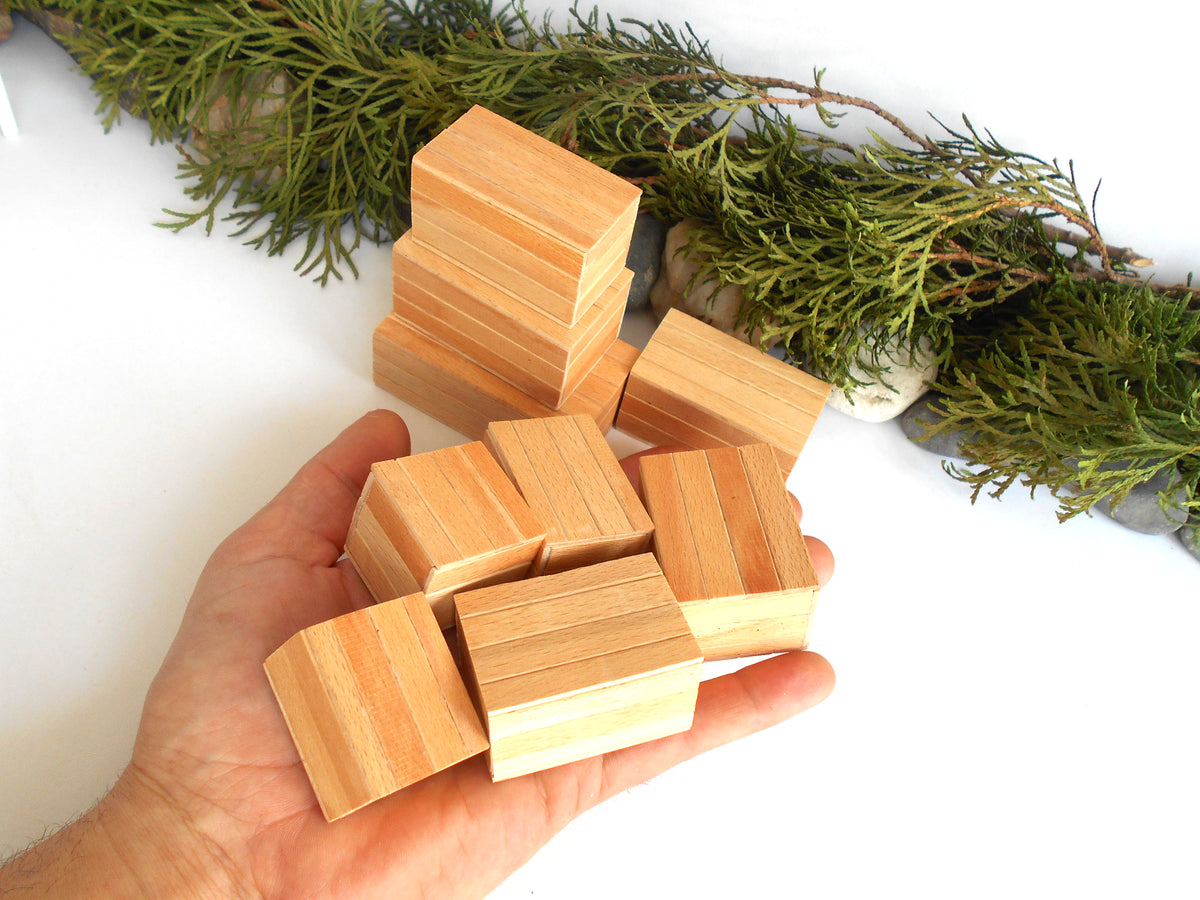 A hand holding several small rectangular wooden miniature boxes with visible wood grain, with more boxes stacked in the background and green foliage decor on a white surface.
