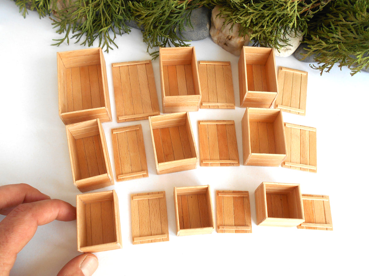 A set of miniature light wood crates and boxes arranged on a white surface, with a person's hand holding one box at the bottom left and greenery at the top.