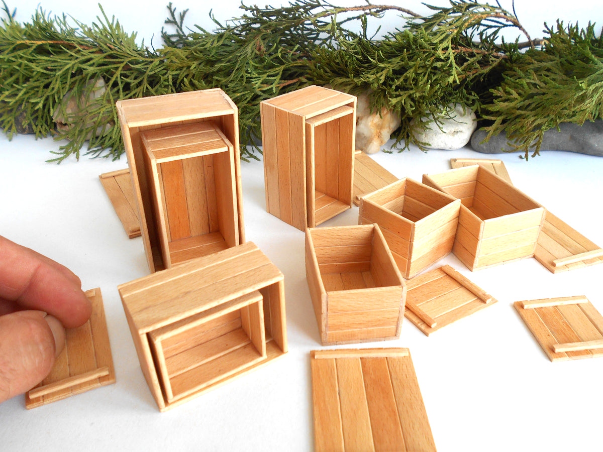 A collection of miniature wooden boxes and crates in various sizes, displayed on a white surface with green pine branches in the background. A human hand is visible on the left, holding one of the small wooden pieces.