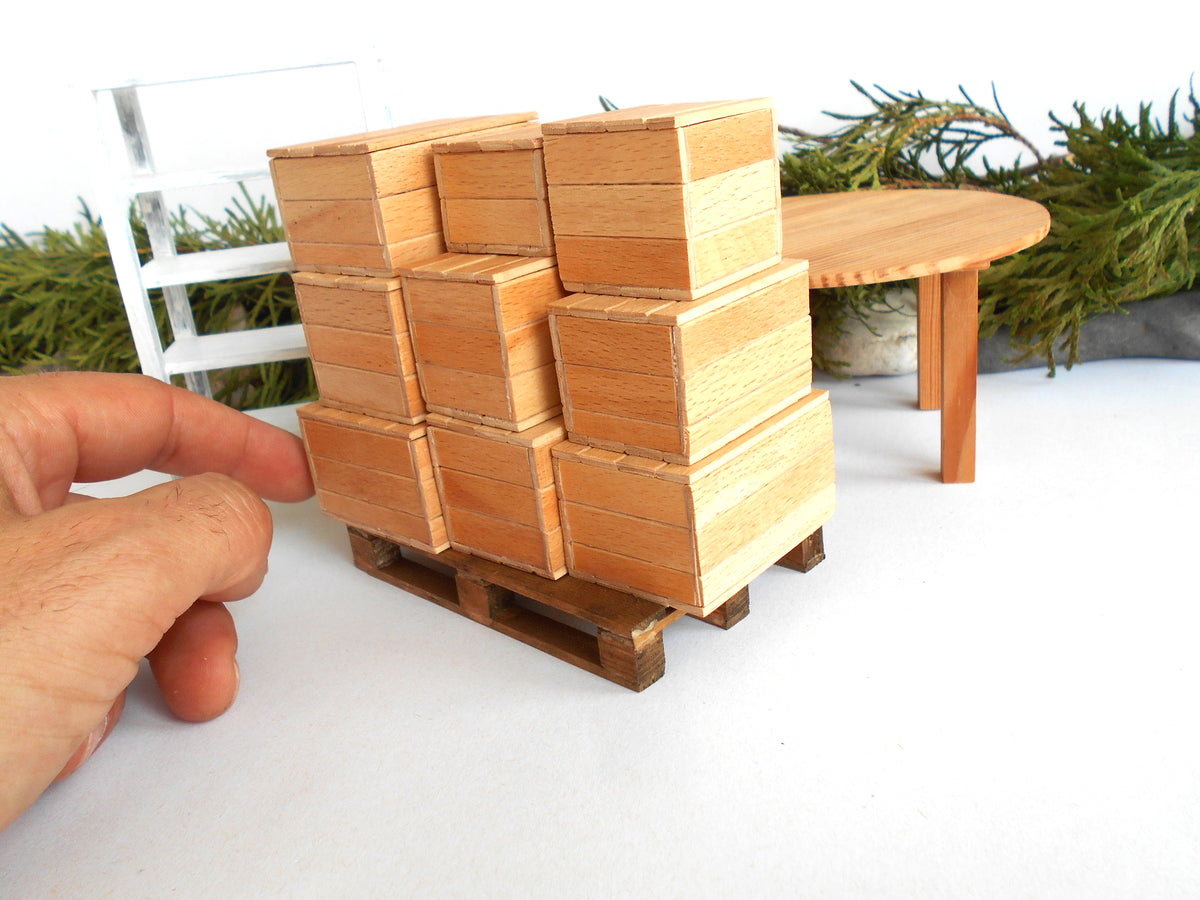 A miniature wooden pallet stacked with small wood miniature boxes , shown next to a human hand for scale, with a miniature table and greenery in the background.