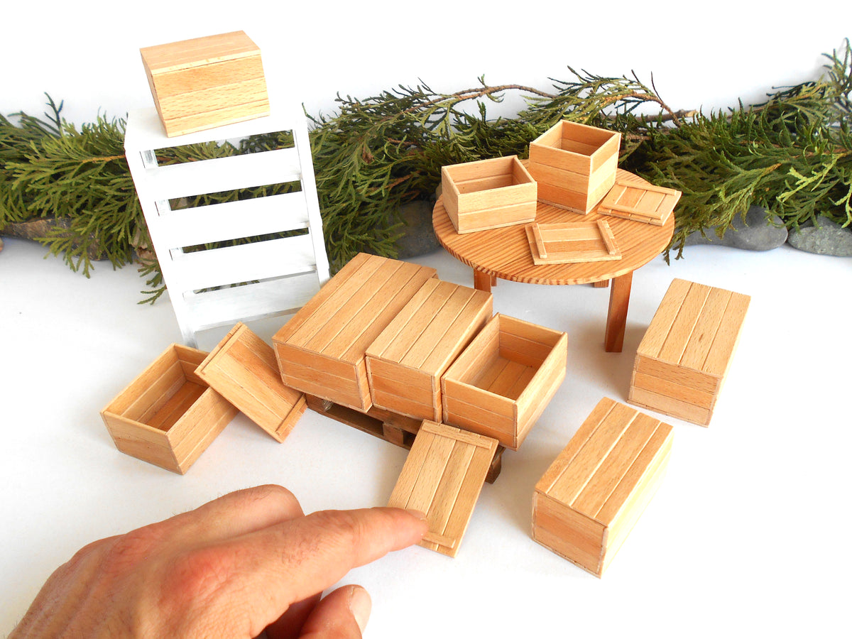 A miniature wooden market scene with small crates, boxes, and a round table, displayed on a white surface with greenery in the background. A human hand is visible in the lower left pointing toward one of the crates.