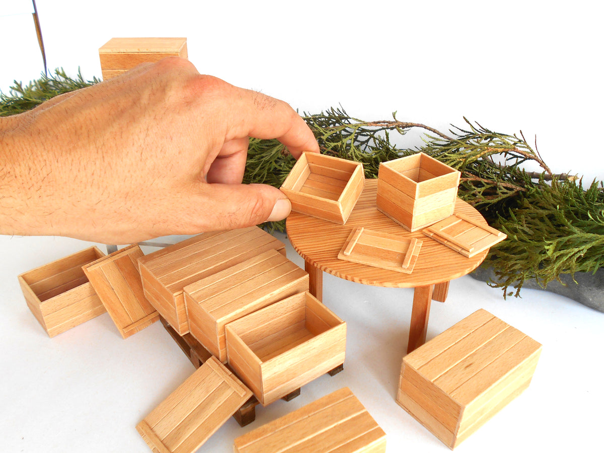 A hand is assembling a miniature wooden table and storage boxes, likely for a diorama or model kit. The set includes light-colored wood grain boxes, drawers, and a round table on a white background with green pine branches as styling props.