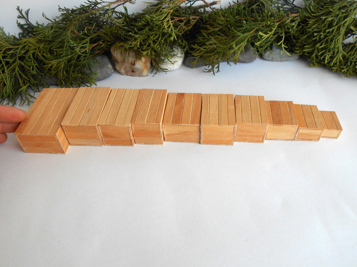 A set of natural wood miniature boxes arranged in a descending size sequence on a white surface, with green foliage and small stones in the background. A hand is visible on the left holding the largest block.