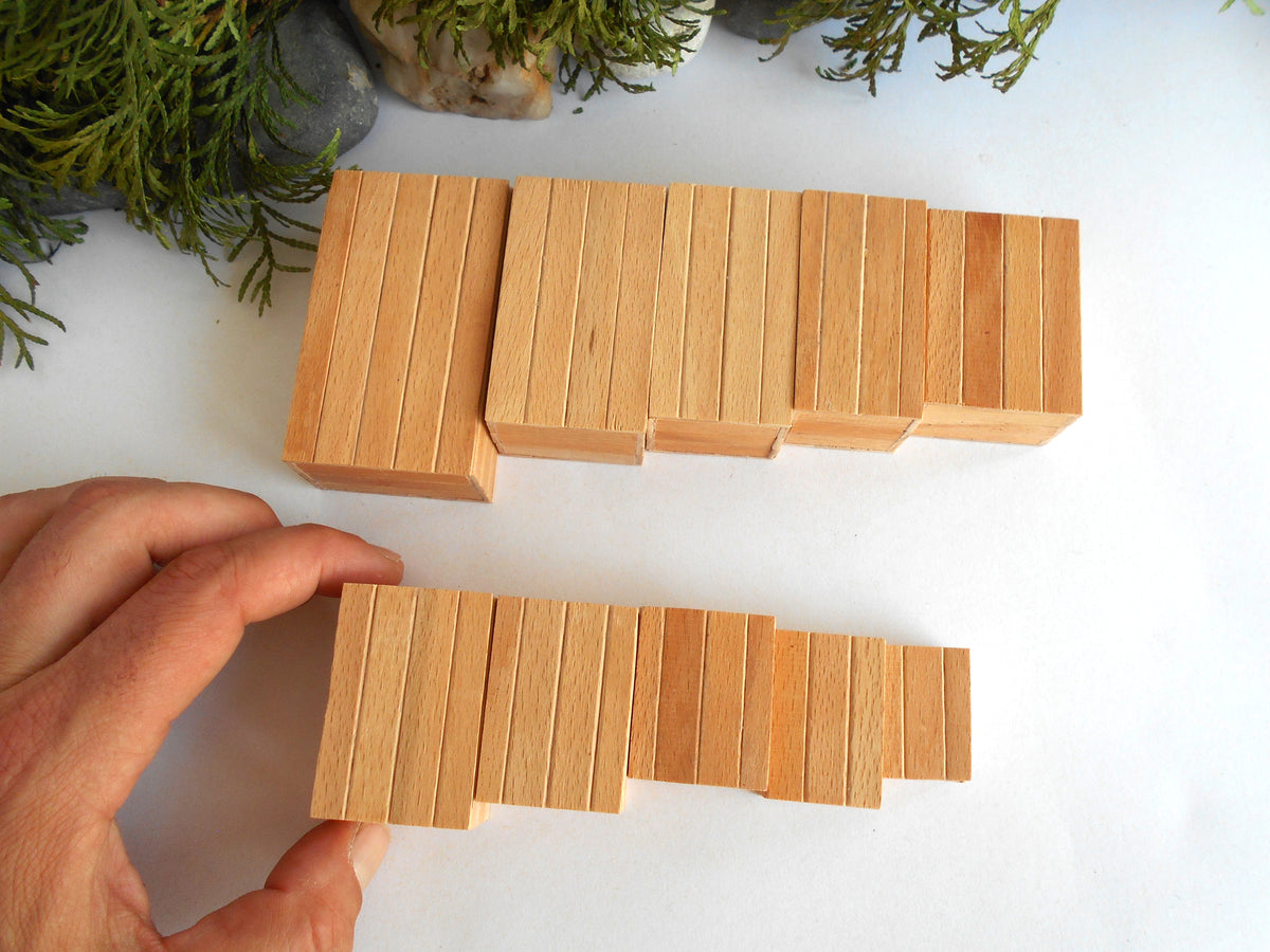 A hand holds a small set of light natural wood miniature boxes , with a larger matching set resting on a white surface in the background. Green foliage decor is visible in the top corners.