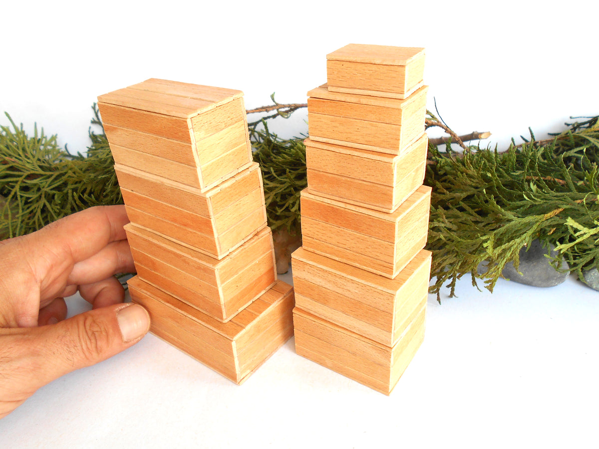 Two stacks of small rectangular wooden miniature boxes in a light natural wood tone, with a hand holding one stack on the left, set against a white background with green foliage props.