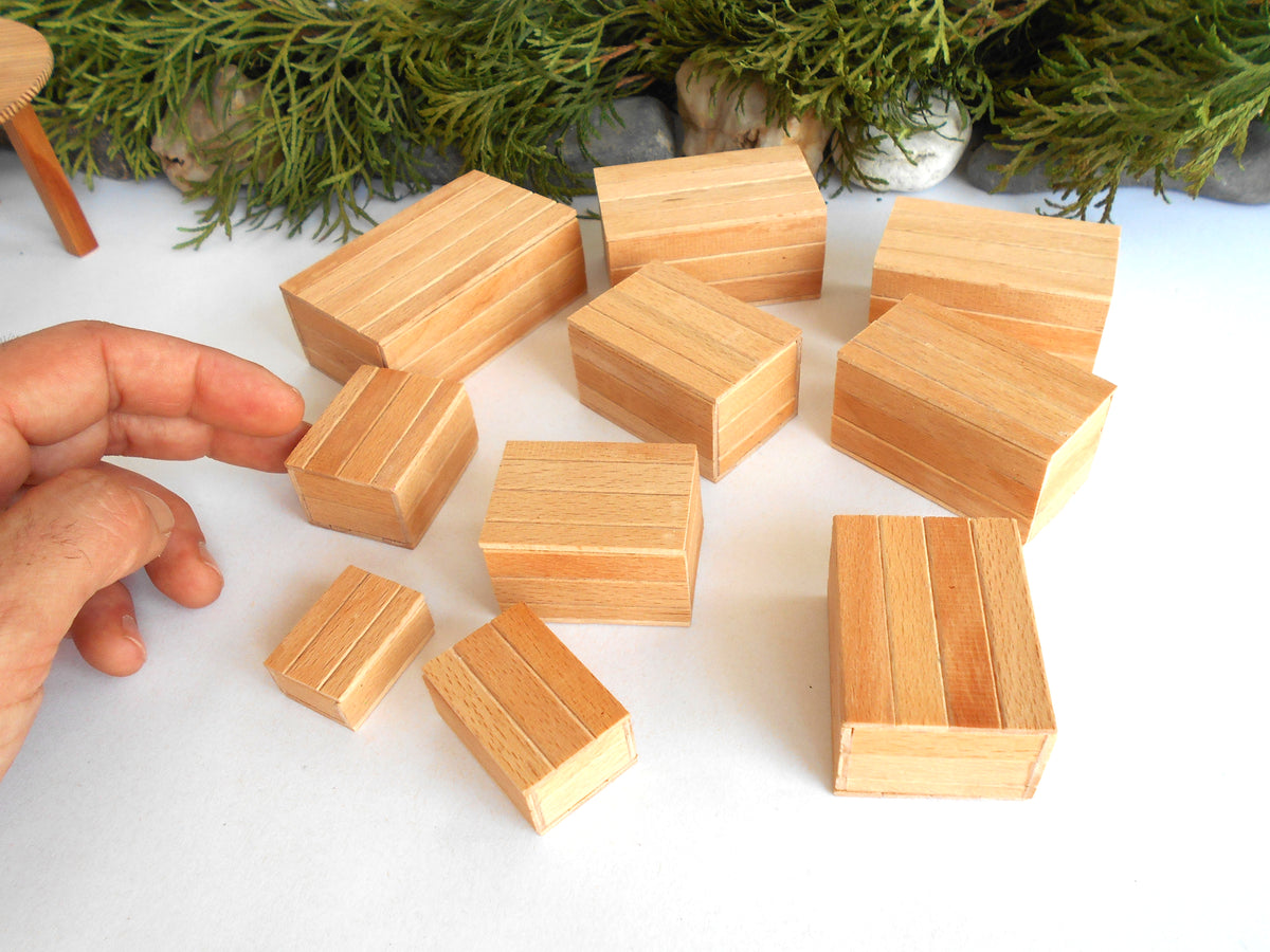 A hand is arranging small rectangular wooden miniature boxes with visible wood grain patterns on a white surface, with green foliage in the background.