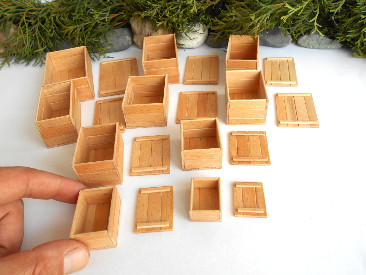 A hand holding a miniature wooden crate next to a collection of small, matching wooden storage boxes and crates arranged on a white surface with green foliage in the background.