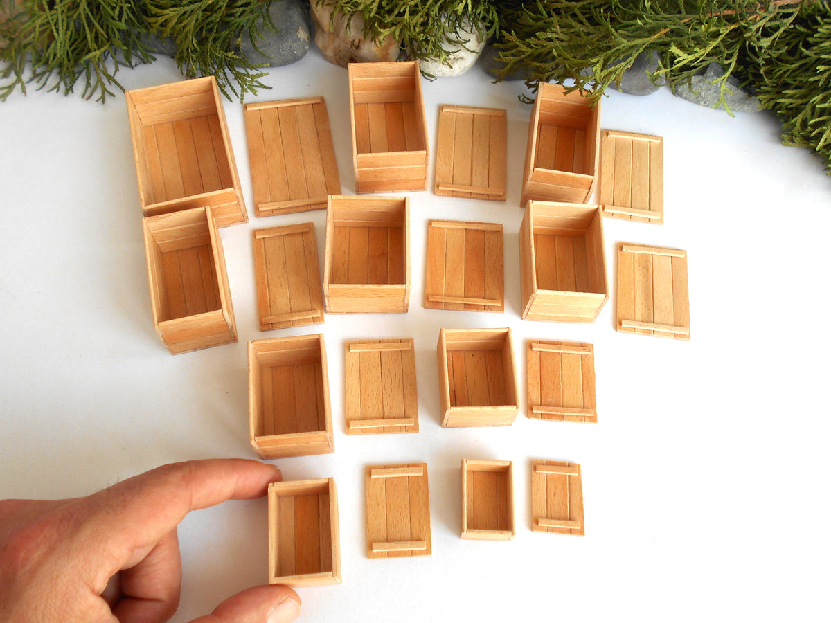 A set of miniature light brown wooden crates arranged on a white surface, with a hand pointing to one of the small crates at the bottom left. Green foliage is visible along the top edge.