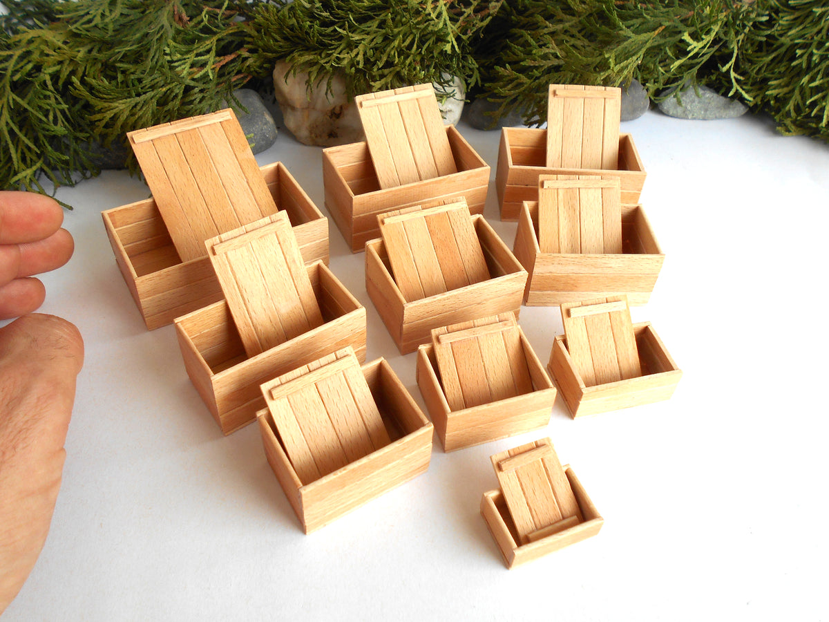 A set of miniature light wood miniature boxes arranged on a white surface, with a hand visible on the left for scale and greenery in the background.