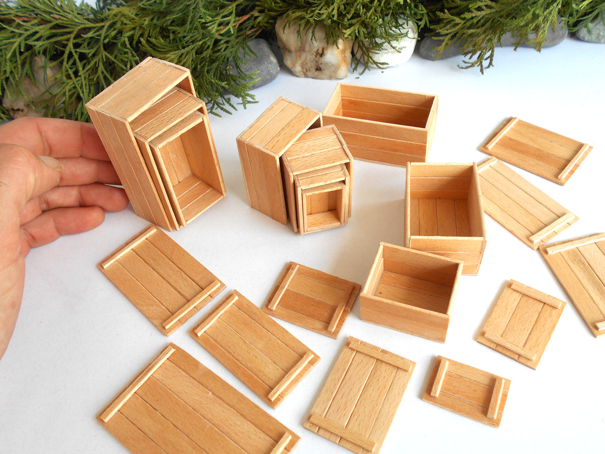 A set of miniature light wood crates and boxes, including some nested sets and flat lids, displayed on a white surface with a hand holding one of the crates on the left.