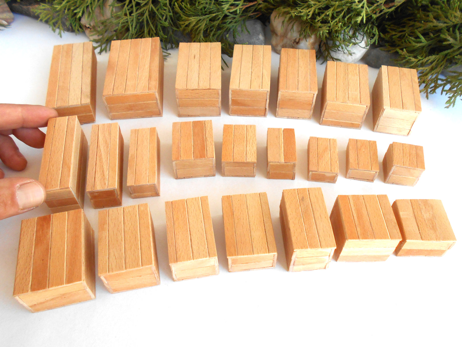 A set of small natural wood miniature boxes or tiles arranged on a white surface, with a hand holding one block on the left and evergreen branches in the background.