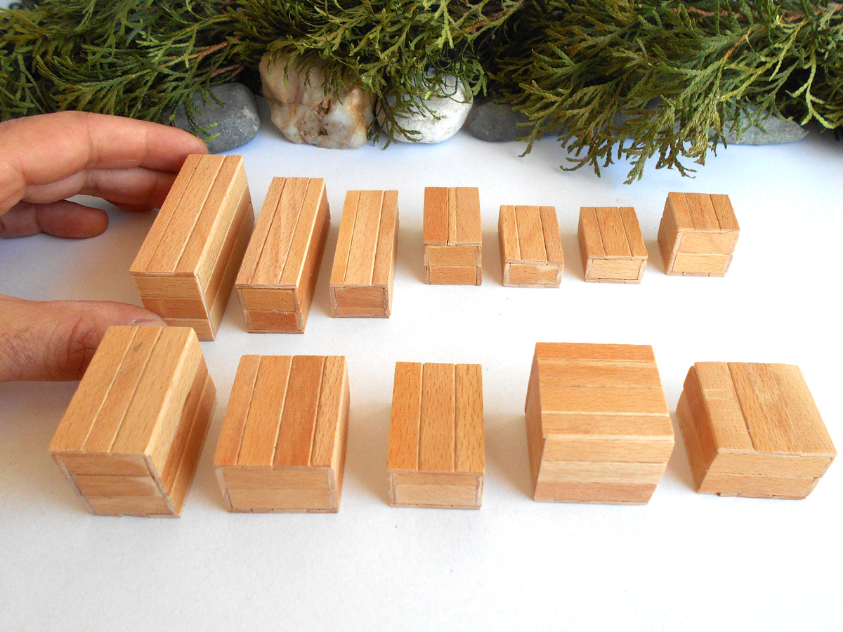 A set of natural wood miniature boxes  in various sizes, arranged on a white surface with a hand holding two of the larger blocks on the left, and green foliage in the background.