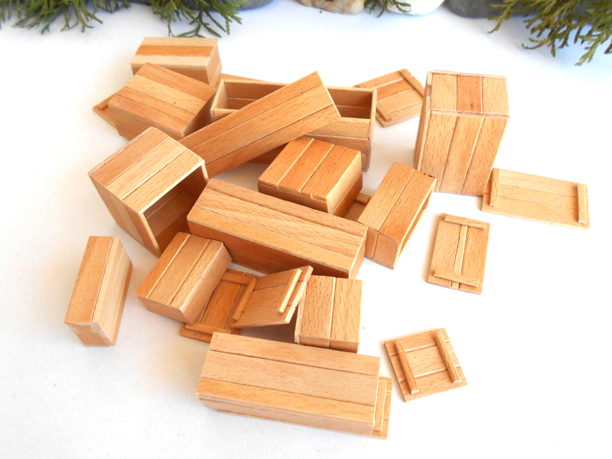 A pile of miniature wooden crates and boxes, likely toy or model building blocks, arranged on a white surface with some green foliage visible at the top edge.