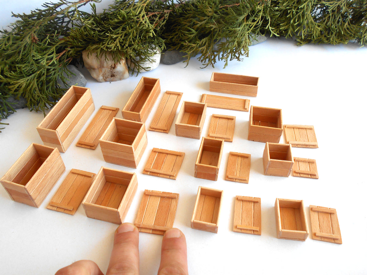 A set of miniature light wood boxes and crates in various sizes, arranged on a white surface with a person's fingers visible at the bottom for scale, and green pine branches at the top.