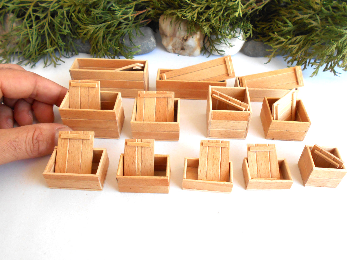 A hand holding a miniature wooden crate next to a collection of small wooden toy or model crates on a white surface with green foliage in the background.