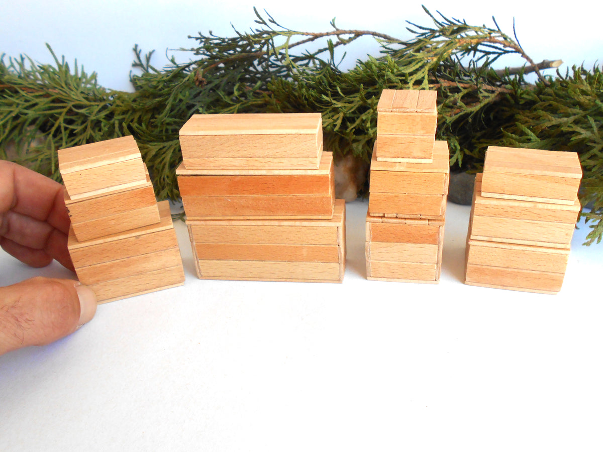 Four small wooden miniature boxes  arranged on a white surface, with a hand holding one stack on the left and evergreen branches in the background.