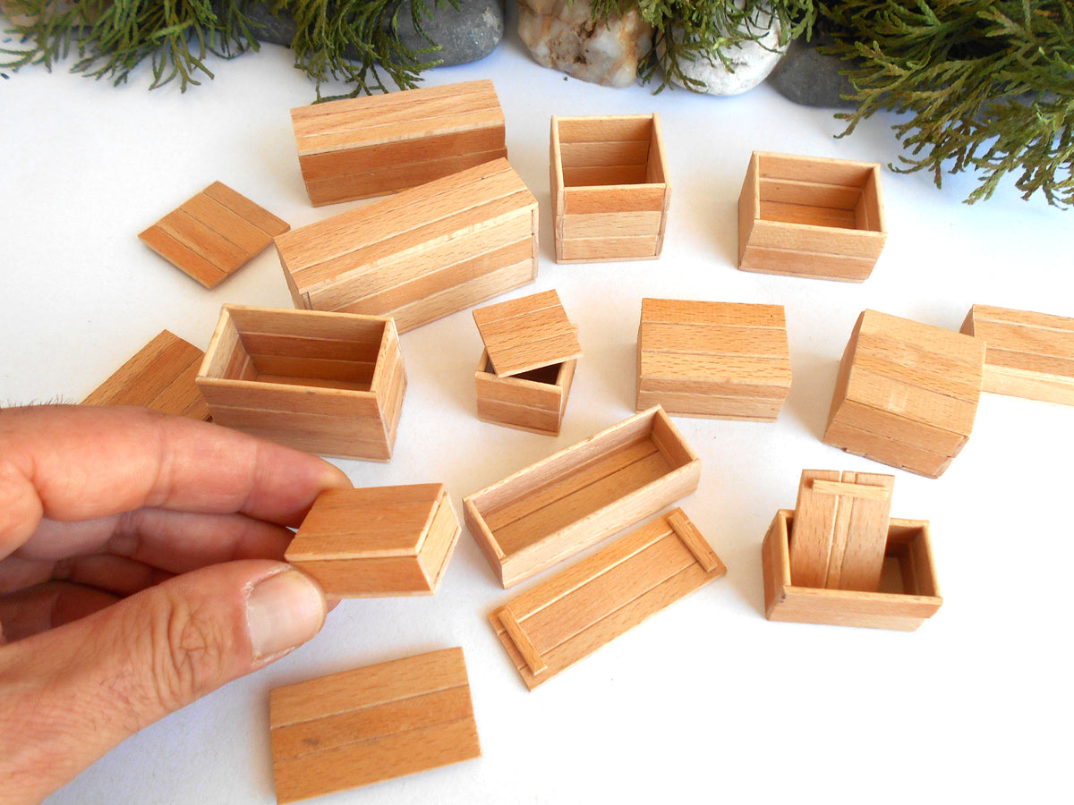 A hand holding a small rectangular wooden box lid, surrounded by multiple matching miniature light wood boxes and compartments on a white surface with decorative greenery in the background.