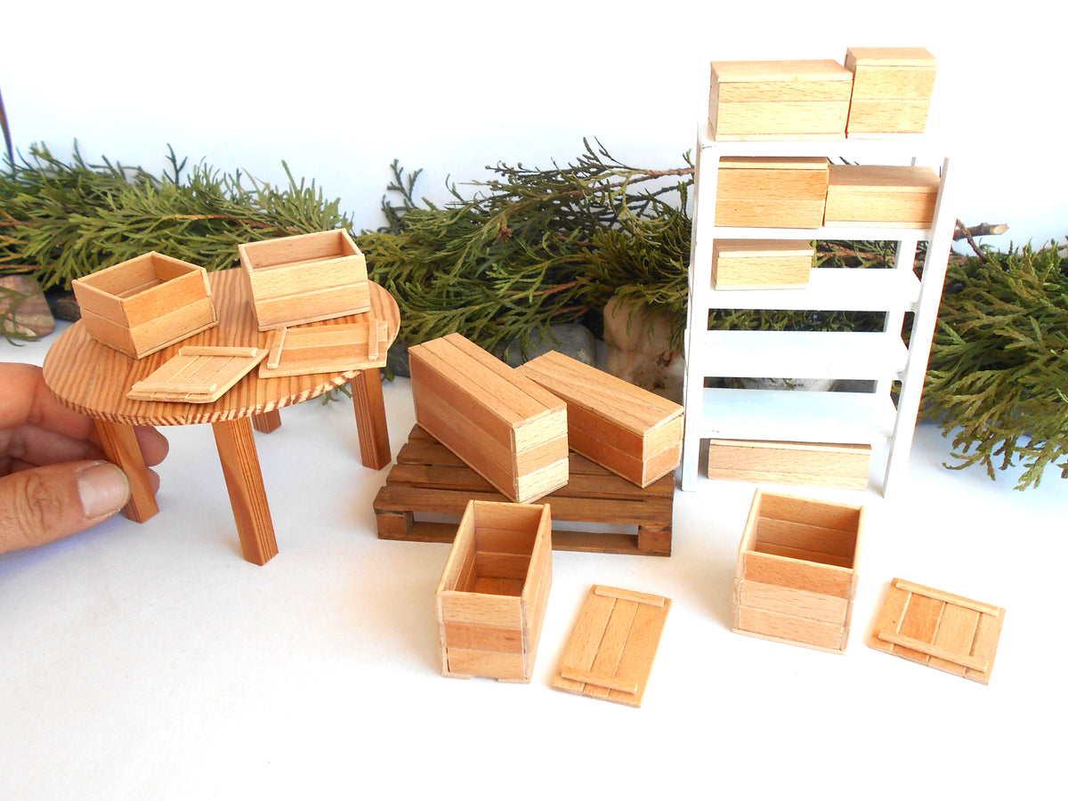 A miniature wooden dollhouse furniture set including a small round table, storage boxes, pallets, and a white shelving unit, with a hand holding one of the wooden crates on the left.