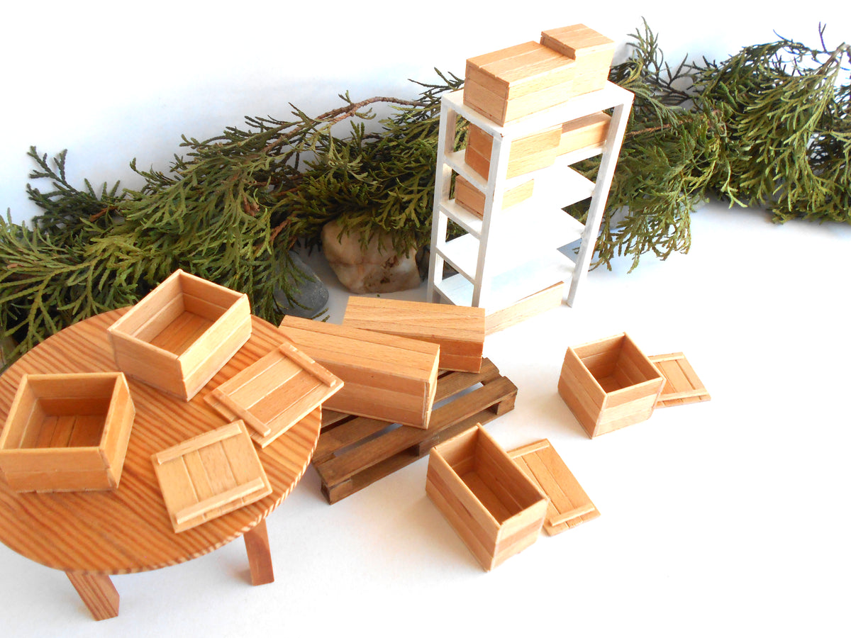 A miniature wooden toy furniture set including a round table, ladder, and various storage boxes, displayed on a white surface with green pine branches in the background.