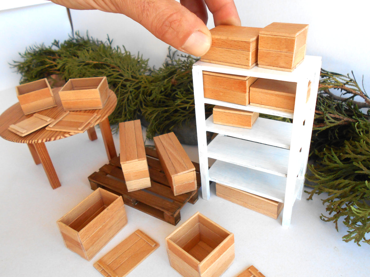 A miniature wooden dollhouse furniture set including a white shelving unit, a round wooden table, crates, and boxes, with a hand placing a wooden box on top of the shelf.