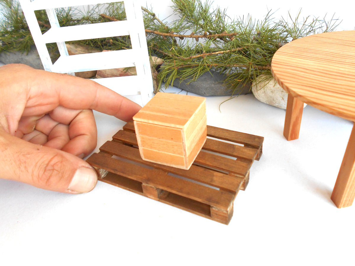 A miniature wooden box sits on a tiny wooden pallet, with a human hand holding it for scale. The scene includes miniature furniture and greenery in the background, suggesting a dollhouse or scale model setup.