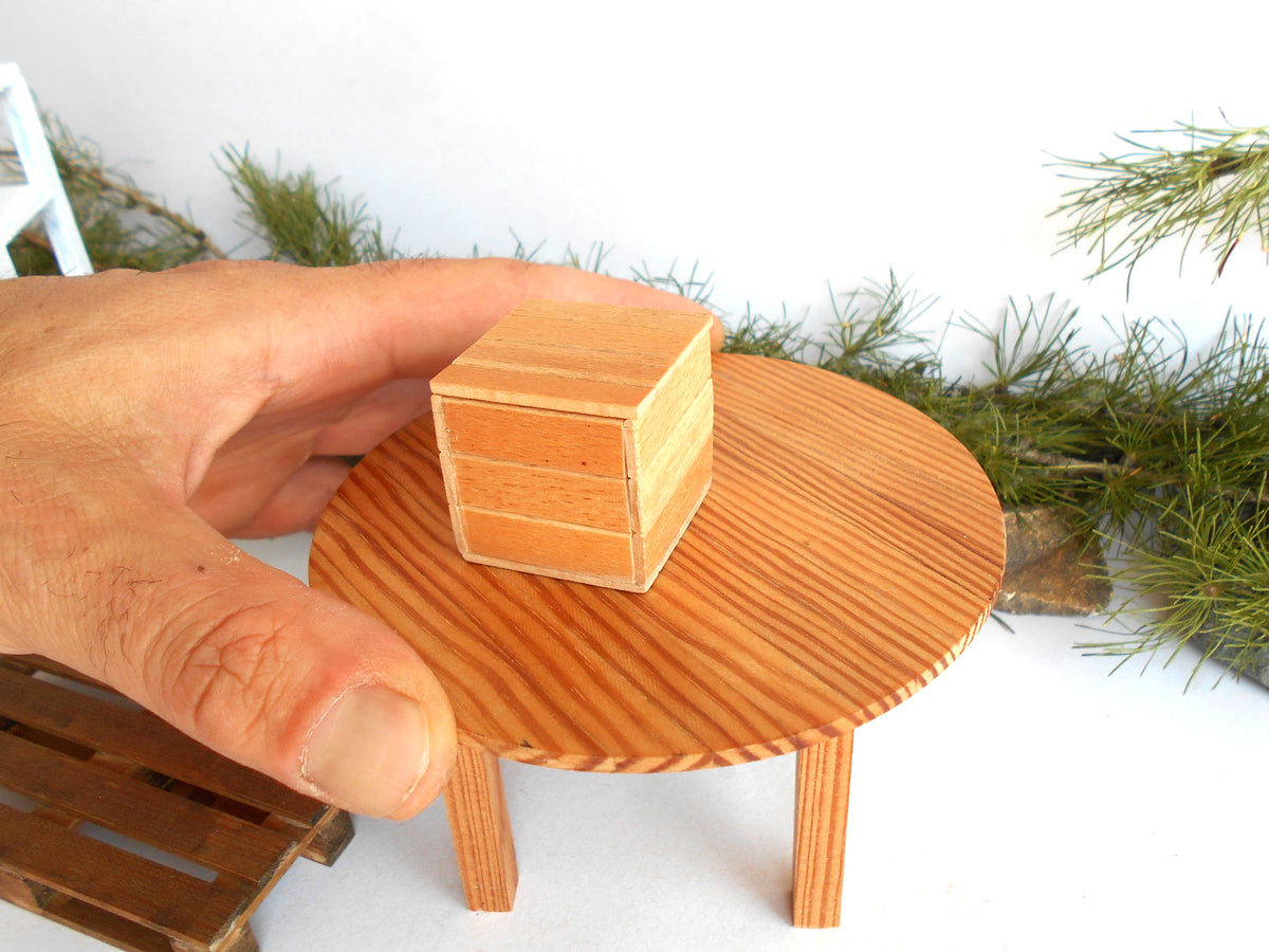 A miniature wooden box with three drawers sits on a small round wooden table, held by a human hand to show scale. The scene is set against a white background with decorative green pine branches.