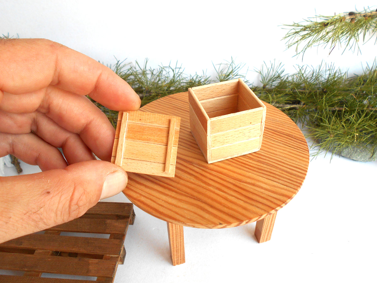 A close-up of a hand holding a small wooden tray next to a matching small wooden box on a miniature round wooden table, with green pine branches in the background.