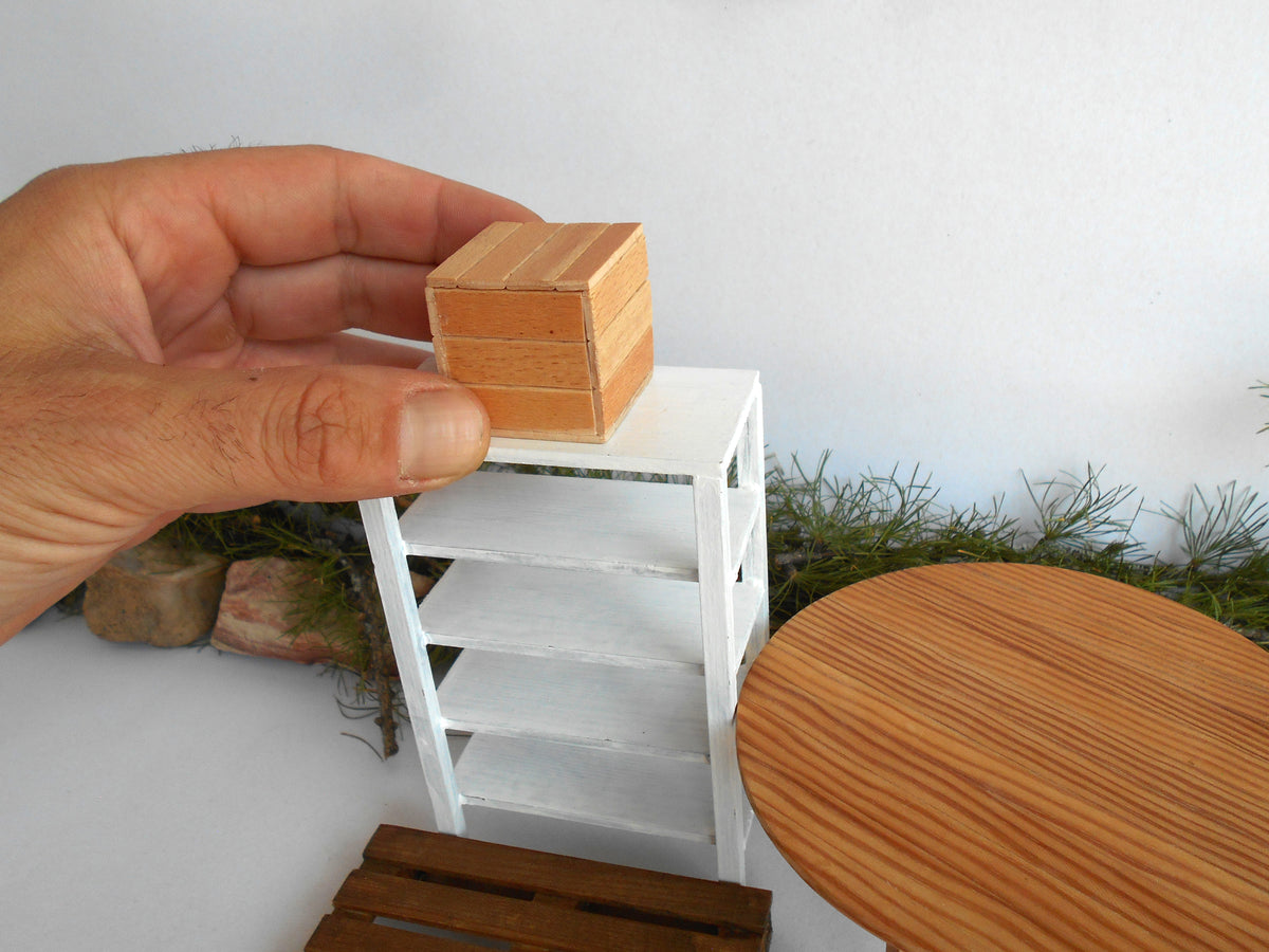 A miniature wooden box with a light natural wood grain finish is being held by a hand and placed on top of a small white miniature shelf. The scene is set against a white background with some miniature greenery and a round wooden table visible.