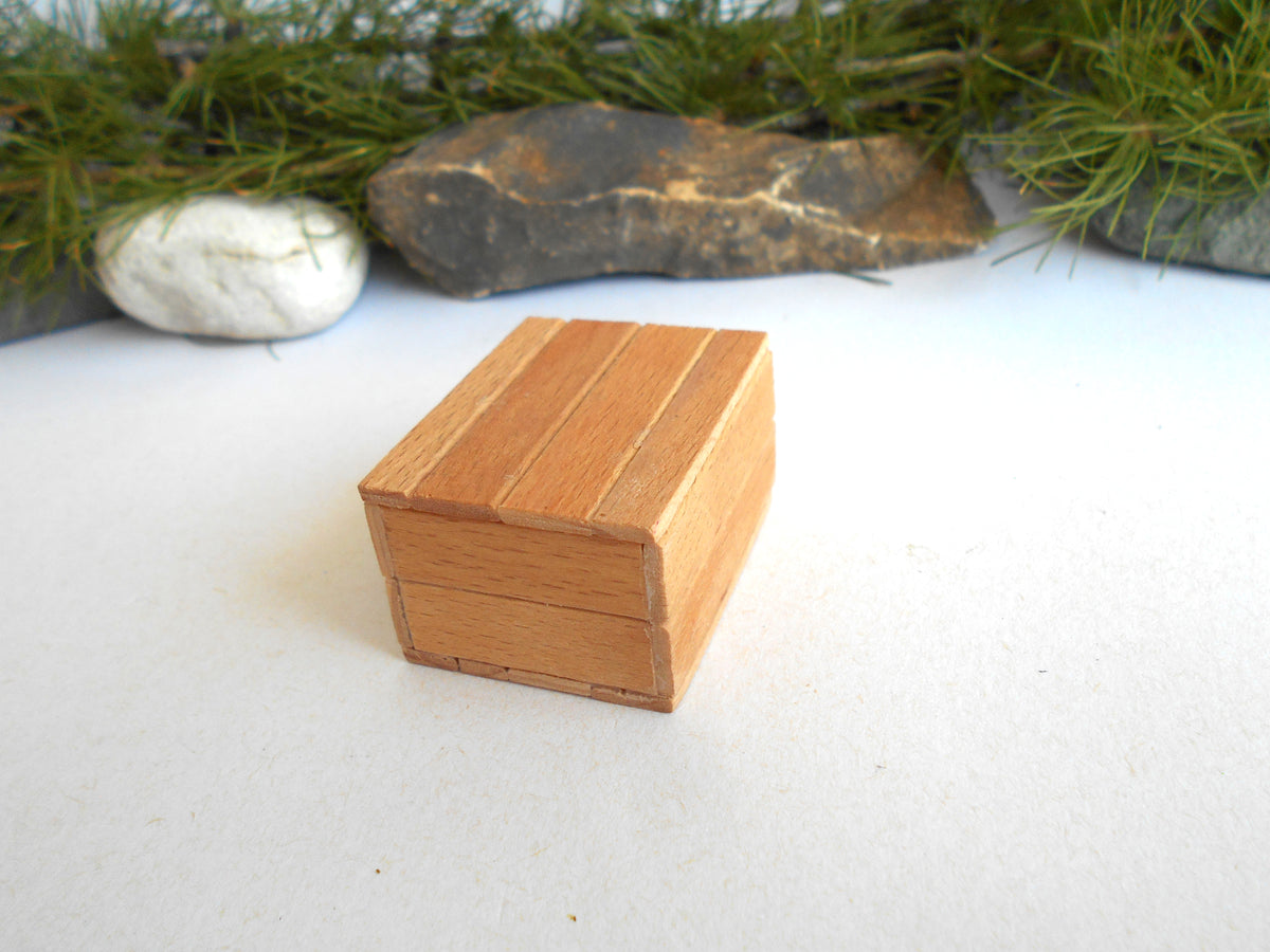 A small rectangular wooden box with a slatted or paneled lid, shown on a white surface with decorative greenery and stones in the background.