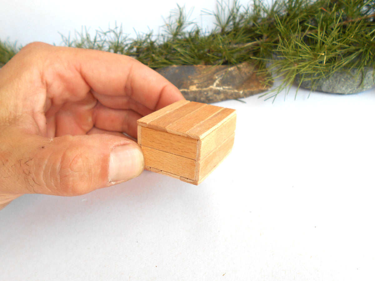 A small rectangular wooden box with a slatted or paneled lid, shown on a white surface with decorative greenery and stones in the 