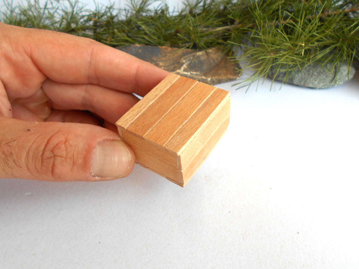 A hand holding a small, square, light brown wooden miniature box with three parallel grooves on its top surface, set against a white background with blurred green pine branches.