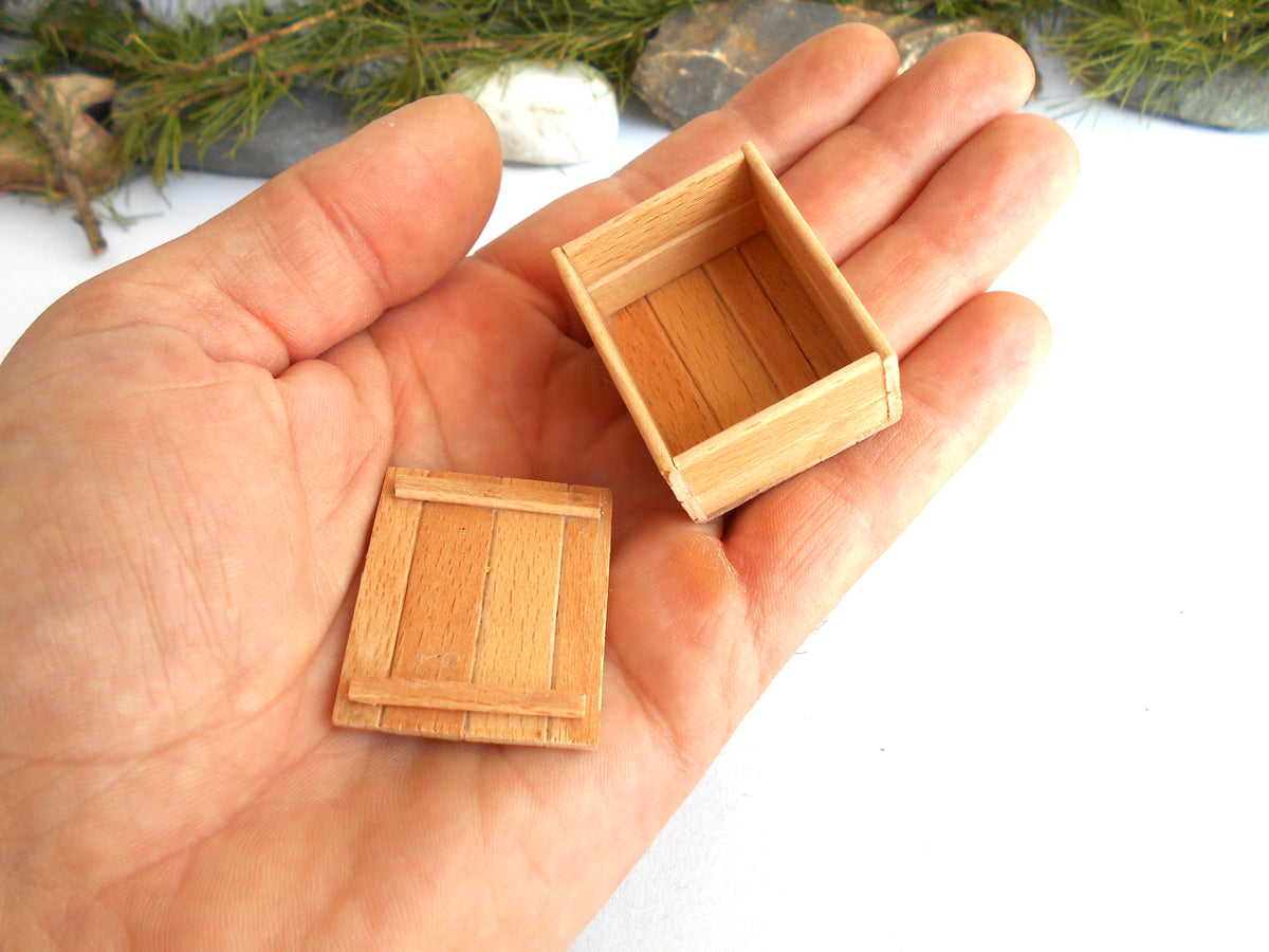 A miniature wooden box and its lid displayed on an open hand, with small green pine branches and stones in the blurred background.
