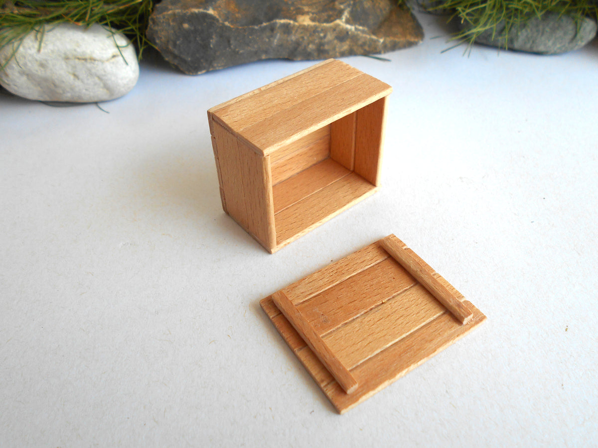 A miniature wooden crate or box with its lid placed in front, photographed on a light surface with small rocks and greenery in the background.