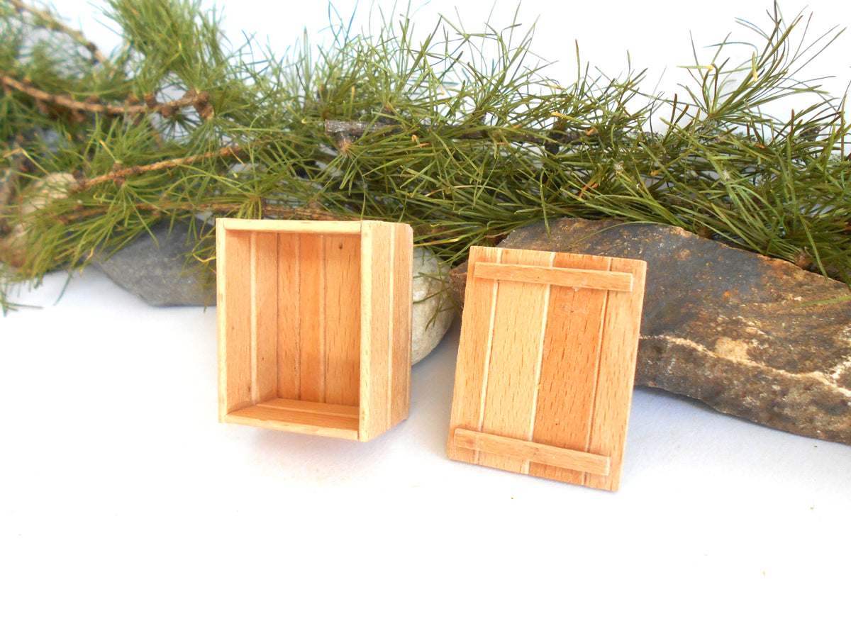 Two miniature light wood crates or boxes displayed on a white surface with pine branches and a rock prop in the background.